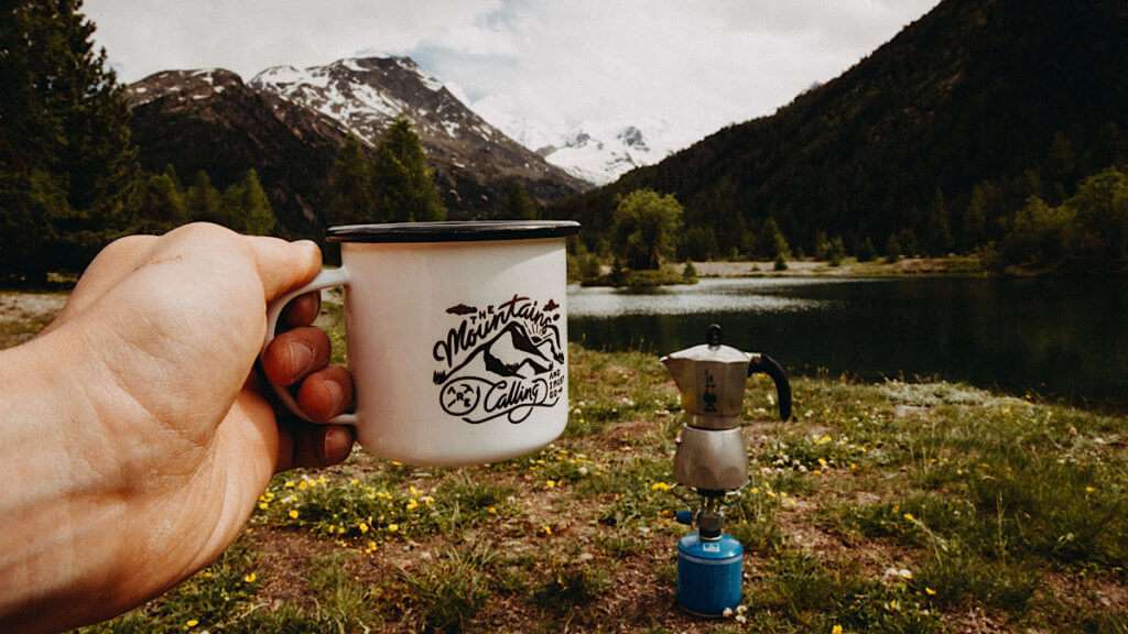 Hand holding a cup in front of a moka pot on s camping stove in front of mountains